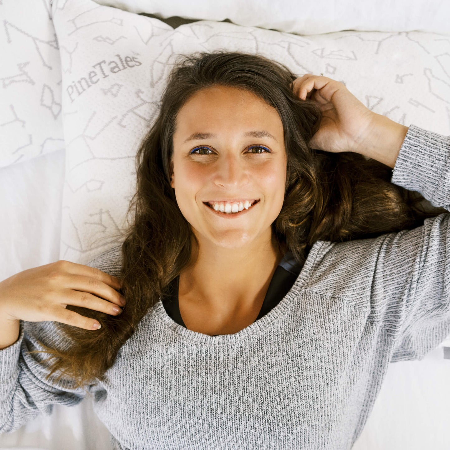 Woman lying on a bed with a 'PineTales' signature buckwheat pillow, smiling and adjusting her hair.